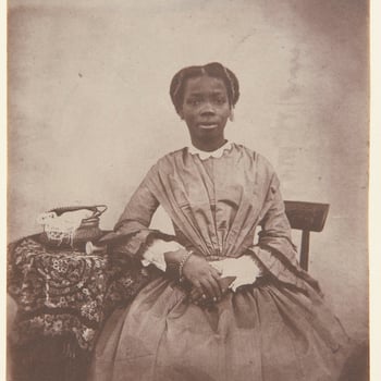 Photograph showing a three-quarters length portrait of Sally [Sarah] Bonetta Forbes, facing the viewer, seated and resting her right arm on a cloth-covered table. The portrait was captioned in the 19th century in ink 'Sally Bonetta Forbes' and dated 1856.