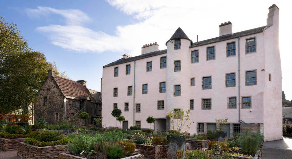 The back of a cream building with a garden in the back. The garden is made of of a serious and raised beds of plants.