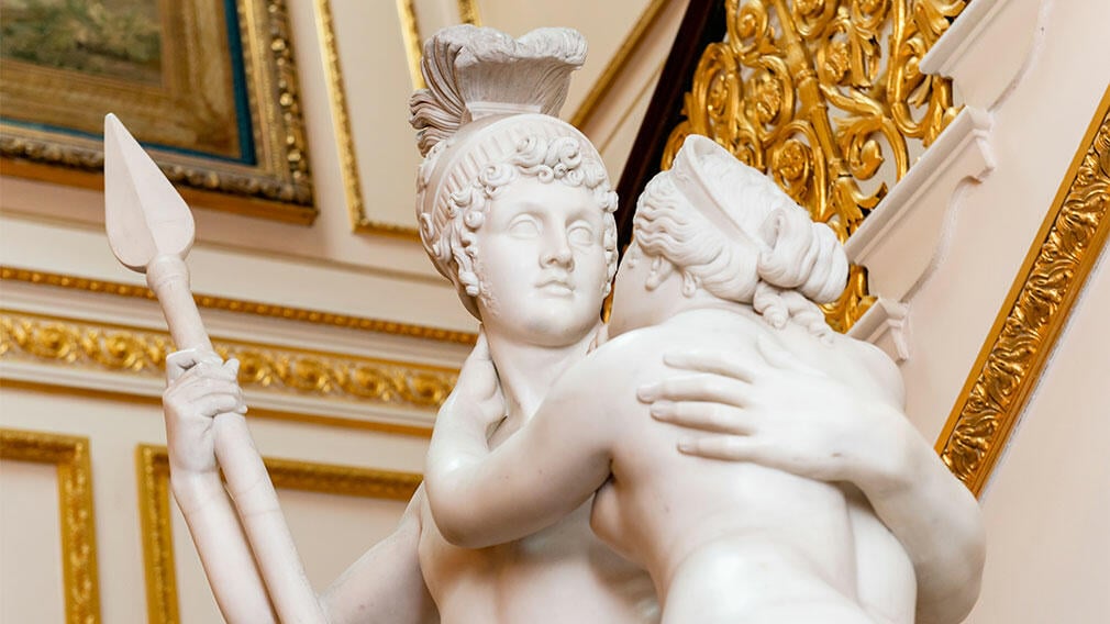 Marble statue of two people embracing with backdrop of palace.