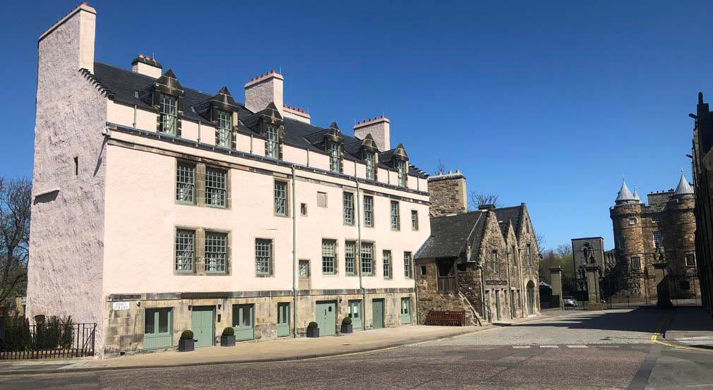 Cream building on the left with a street in front. To the right are the gate and entrance to the Palace of Holyroodhouse.