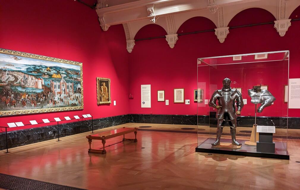 A gallery space with red walls, wooden floor, a wooden bench and exhibits including a suit of armour and paintings.