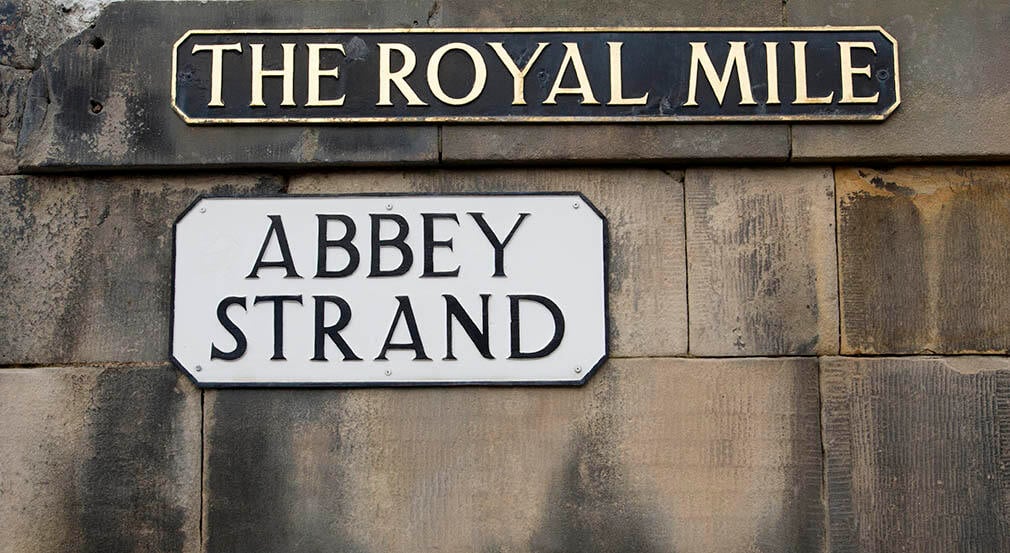 A image of a brick wall with a street sign with the text 'The Royal Mile' and another sign with the text 'Abbey Strand'.