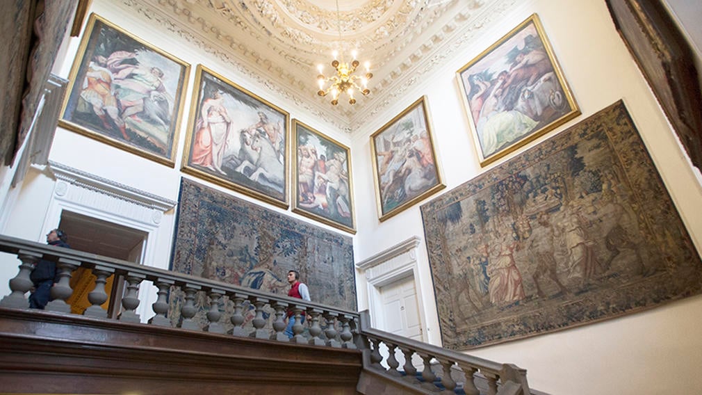 A man walks along a landing of a stone staircase. The walls are filled with tapestries and paintings.